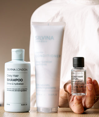 Three Silvina London grey hair products are arranged on a light wood surface: a grey shampoo bottle, a white conditioner tube, and a hand holding a small grey hair oil bottle. A person in a white top is blurred in the background.
