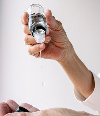 A close-up of a person's hands holding a small clear bottle and dispensing a liquid dropper onto the other hand against a plain light background.