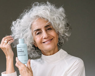 Silvina with grey hair holding a bottle of shampoo, surrounded by various hair care products on a table.