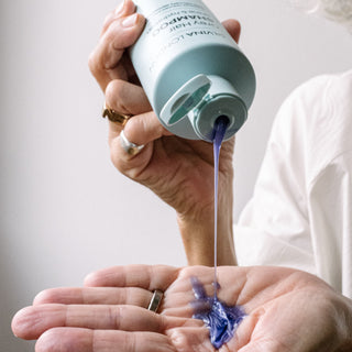 Person pouring purple shampoo from a bottle into their hand against a neutral background